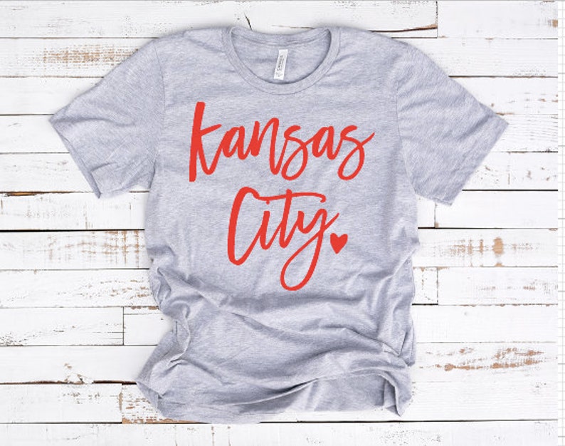 Op de afbeelding: Een lichtgrijs t-shirt met de tekst "Kansas City" in rode letters. Onder het woord "City" staat een klein rood hartje. Het shirt is gemaakt van een zacht materiaal en heeft korte mouwen.
