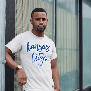 Op de afbeelding: Wit T-shirt met de woorden "Kansas City" in blauw script. Het shirt heeft een klein blauw hartje onder het woord "City". De persoon draagt een horloge met een bruine leren band.