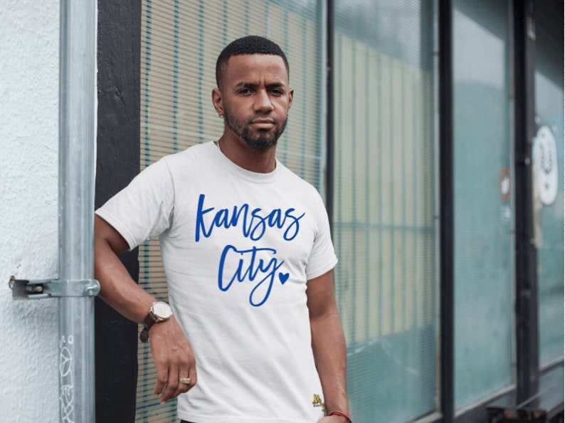 Op de afbeelding: Wit T-shirt met de woorden "Kansas City" in blauw script. Het shirt heeft een klein blauw hartje onder het woord "City". De persoon draagt een horloge met een bruine leren band.