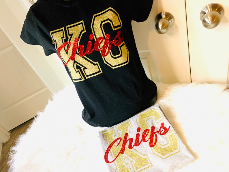 Op de afbeelding: Twee T-shirts met de tekst "KC Chiefs" in gouden glitterletters. Het bovenste shirt is zwart en het onderste shirt is grijs. Het woord "Chiefs" is in rode glitterletters.