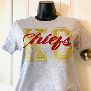Op de afbeelding: Een grijs T-shirt met een gouden en rood glitterontwerp met de tekst "KC Chiefs".