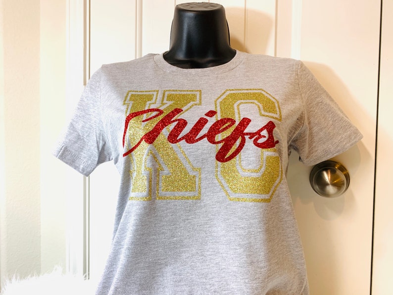 Op de afbeelding: Een grijs T-shirt met een gouden en rood glitterontwerp met de tekst "KC Chiefs".
