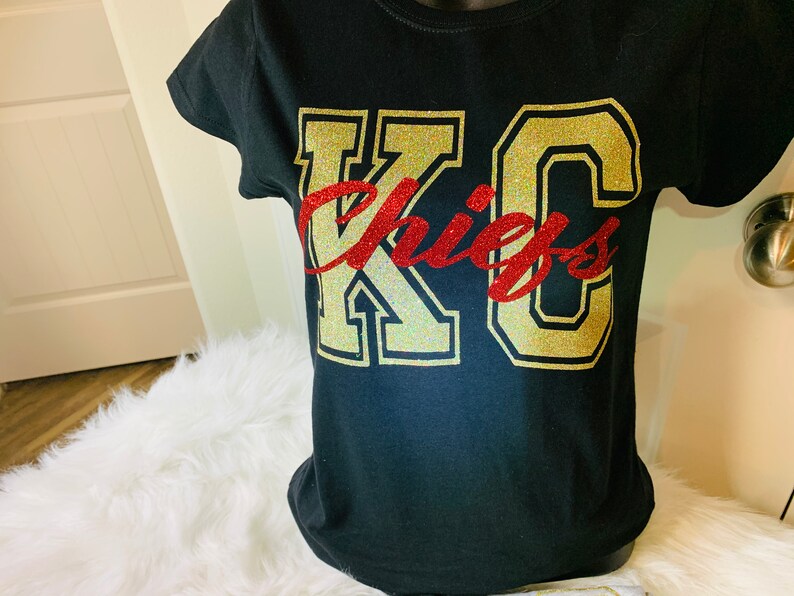 Op de afbeelding: Zwart T-shirt met gouden glitterletters met de tekst "KC Chiefs" in een lettertype in varsity-stijl.