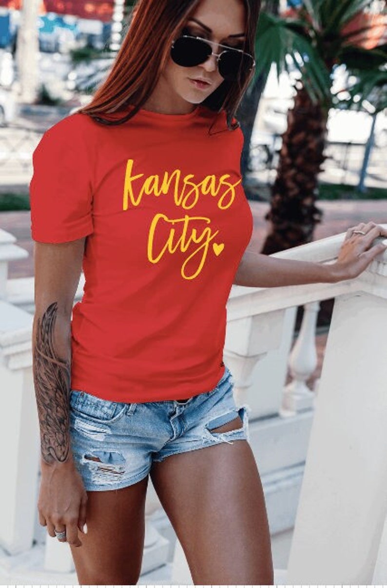 Op de afbeelding: Een rood t-shirt met de woorden "Kansas City" in gele letters. Het shirt wordt gecombineerd met een spijkerbroek. De foto is buiten gemaakt.