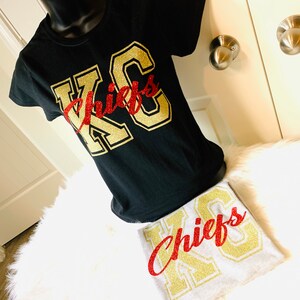 Op de afbeelding: Twee T-shirts met de tekst "KC Chiefs" in gouden glitterletters. Een shirt is zwart met rode glitterletters en het andere is grijs met rode glitterletters.