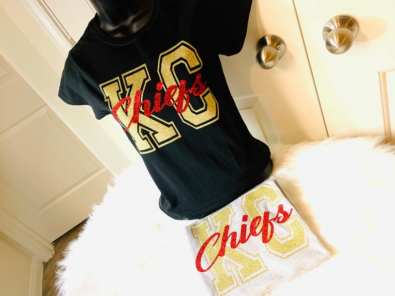 Op de afbeelding: Twee T-shirts met de tekst "KC Chiefs" in gouden glitterletters. Een shirt is zwart met rode glitterletters en het andere is grijs met rode glitterletters.