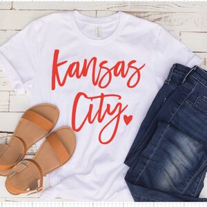 Op de afbeelding: Wit t-shirt met de woorden "Kansas City" in rode letters en een rood hart. Het shirt wordt gecombineerd met een blauwe jeans en bruine sandalen. De sandalen hebben een platte zool en een enkelbandje.