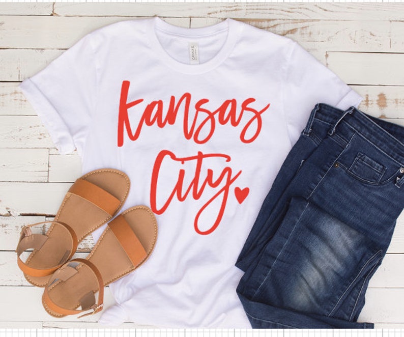 Op de afbeelding: Wit t-shirt met de woorden "Kansas City" in rode letters en een rood hart. Het shirt wordt gecombineerd met een blauwe jeans en bruine sandalen. De sandalen hebben een platte zool en een enkelbandje.