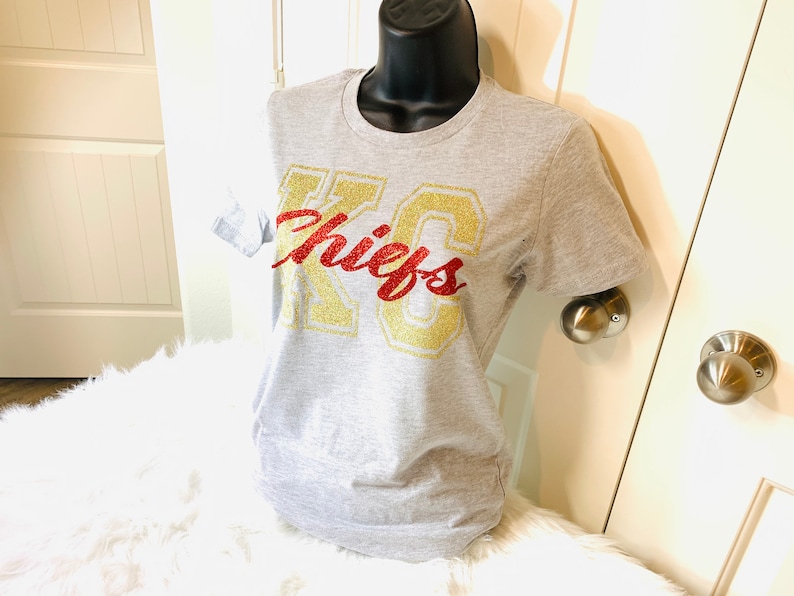 Op de afbeelding: Een grijs T-shirt met een gouden glitter "KC Chiefs" afbeelding.