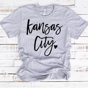 Op de afbeelding: Lichtgrijs t-shirt met de woorden "Kansas City" in zwart script. Een klein zwart hartje staat onder "City". Het shirt is gemaakt van een zacht materiaal en heeft een ronde hals.