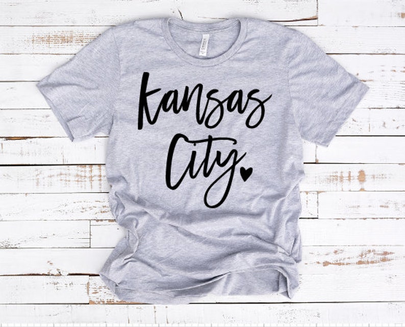 Op de afbeelding: Lichtgrijs t-shirt met de woorden "Kansas City" in zwart script. Een klein zwart hartje staat onder "City". Het shirt is gemaakt van een zacht materiaal en heeft een ronde hals.