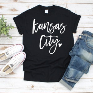 Op de afbeelding: Zwart T-shirt met de woorden "Kansas City" in een wit scriptlettertype en een klein hartje. Het shirt wordt gecombineerd met een blauwe jeans en witte sneakers. Een kleine groene plant staat ook in de afbeelding.