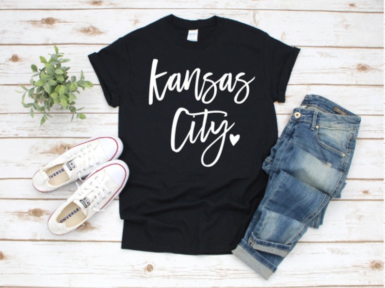 Op de afbeelding: Zwart T-shirt met de woorden "Kansas City" in een wit scriptlettertype en een klein hartje. Het shirt wordt gecombineerd met een blauwe jeans en witte sneakers. Een kleine groene plant staat ook in de afbeelding.