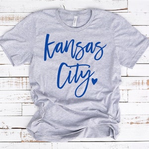 Op de afbeelding: Een lichtgrijs t-shirt met de woorden "Kansas City" in blauw script, met een blauw hartje dat de punt boven de "i" in "City" vervangt. Het shirt is gemaakt van een zacht materiaal.