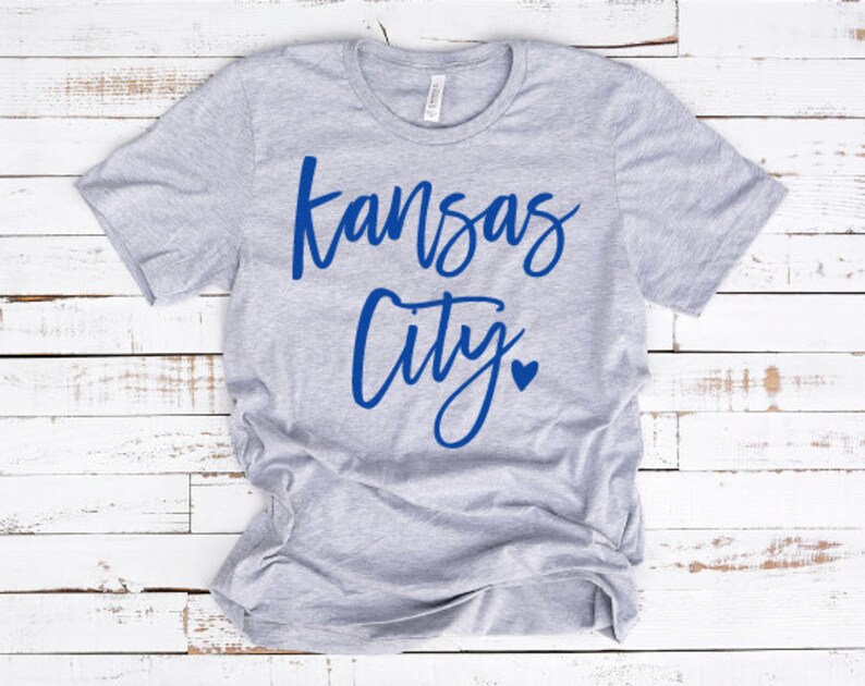 Op de afbeelding: Een lichtgrijs t-shirt met de woorden "Kansas City" in blauw script, met een blauw hartje dat de punt boven de "i" in "City" vervangt. Het shirt is gemaakt van een zacht materiaal.