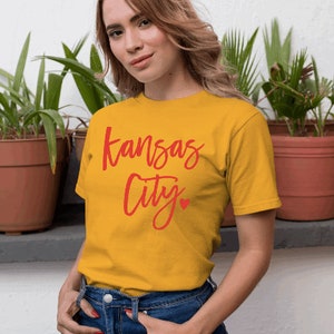 Op de afbeelding: Mosterdgeel T-shirt met de woorden "Kansas City" in rode letters en een klein rood hart. Het shirt wordt gecombineerd met een blauwe spijkerbroek. Het model staat voor een witte muur met potplanten.