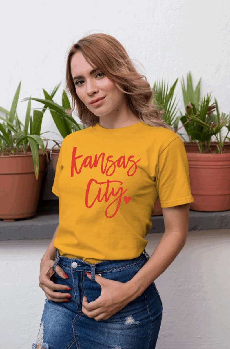 Op de afbeelding: Mosterdgeel T-shirt met de woorden "Kansas City" in rode letters en een klein rood hart. Het shirt wordt gecombineerd met een blauwe spijkerbroek. Het model staat voor een witte muur met potplanten.