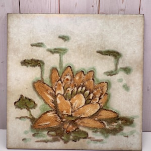 May include: Square ceramic tile featuring a detailed illustration of a lotus flower in shades of orange and brown, surrounded by green lily pads and stems. The background has a mottled, off-white appearance, creating a vintage aesthetic.