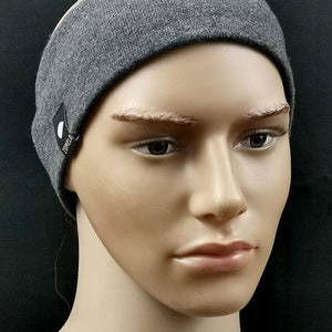 Peut inclure: Un bandeau gris avec une petite étiquette noire avec du texte blanc et un motif circulaire. Le bandeau est fait d'un matériau tricoté doux et est porté sur une tête de mannequin. Le fond est noir uni.