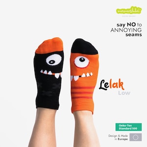 Könnte beinhalten: Zwei lustige Socken, eine schwarze und eine orangefarbene, jeweils mit einem Monstergesicht, großen Augen und Zähnen. Die Socken sind mit "Lelak Low" beschriftet, und der Text "say NO to ANNOYING seams" ist ebenfalls sichtbar.