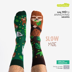 Könnte beinhalten: Ein Paar bunte Socken mit einem Faultier-Motiv. Die Socken sind grün und braun mit einem Muster aus Faultieren und Blättern. Der Text "say NO to ANNOYING seams" ist auf den Socken aufgedruckt. Der Text "SLOW MOE" ist unter den Socken aufgedruckt. Die Socken werden in Europa hergestellt und sind Oeko-Tex Standard 100 zertifiziert.