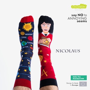 Könnte beinhalten: Ein Paar bunte Socken. Eine Socke hat einen roten Hintergrund mit einer Cartoon-Illustration einer Person mit einem roten Shirt und einem Buch. Die andere Socke hat einen blauen Hintergrund mit einer Grafik von Planeten und Sternen. Die Socken haben den Text "say NO to ANNOYING seams" und "NICOLAUS" aufgedruckt. Die Socken sind aus Oeko-Tex Standard 100 zertifizierten Materialien hergestellt und wurden in Europa entworfen und hergestellt.
