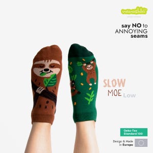 Op de afbeelding: Een paar novelty sokken met luiaardontwerpen. Eén sok is bruin met een luiaard op een tak, de andere is groen met luiaards en bladeren. De tekst op de sokken luidt "SLOW MOE Low" en "say NO to ANNOYING seams."