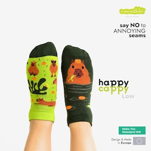 Może przedstawiać: Para zielonych i żółtych skarpet z ilustracjami kapibar. Jedna skarpetka przedstawia kapibary w stawie, druga na lądzie. Na skarpetkach widnieje napis "happy cappy" i "say NO to ANNOYING seams."