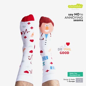 Op de afbeelding: Witte sokken met rode details, versierd met medische symbolen, waaronder harten, kruisen en een dokter illustratie. De sokken tonen de tekst "say NO to ANNOYING seams" en "DR PHIL GOOD". Ze zijn voorzien van het label "Oeko-Tex Standard 100".
