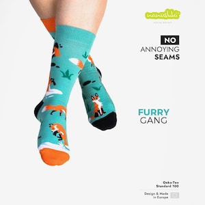 Könnte beinhalten: Türkisfarbene Socken mit orangefarbenen Füchsen, weißen Wolken und grünem Laub. Die Socken haben schwarze Fersen und Zehen sowie orangefarbene Bündchen. Auf den Socken steht "FURRY GANG" und "NO ANNOYING SEAMS". Sie sind mit "Oeko-Tex Standard 100" und "Design & Made in Europe" gekennzeichnet.