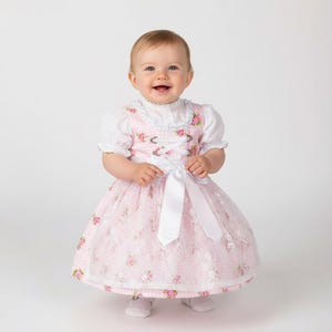 Festliches Baby Dirndl aus Baumwolle mit einem Blumen Muster in rosa-weiß mit einer Schürze aus Spitze.