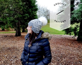 May include: A gray and white crochet beanie with a white pom pom. The beanie is being worn by a person in a black jacket, standing in a forest.