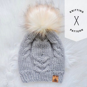 May include: Gray knitted beanie with a large white faux fur pom pom. The beanie has a cable knit pattern and a small brown leather tag with the words "Made with Love" on it.