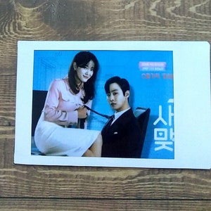 Pode incluir: Uma foto de um homem e uma mulher em uma cena do drama coreano "Love (ft. Marriage and Divorce)". O homem est&aacute; vestindo um terno preto e a mulher est&aacute; vestindo uma camisa rosa e uma saia branca. O texto "사랑은" &eacute; vis&iacute;vel na imagem.