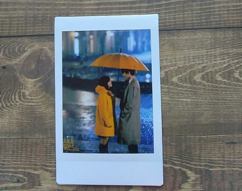 Pode incluir: Um casal est&aacute; debaixo de um guarda-chuva amarelo na chuva. A mulher est&aacute; usando um casaco amarelo e o homem est&aacute; usando um casaco cinza. O texto "시맨틱" &eacute; vis&iacute;vel no canto inferior esquerdo da imagem.