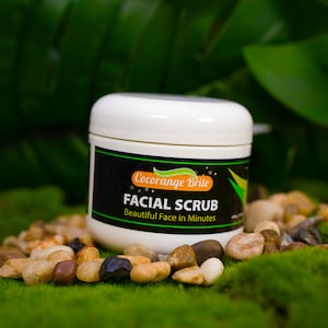 Puede incluir: Un tarro blanco con una etiqueta negra que dice "Cocorange Brile Facial Scrub Beautiful Face in Minutes". El tarro está sentado en una cama de musgo verde y pequeñas piedras marrones.