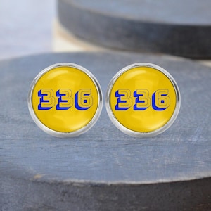 May include: Two circular silver earrings. Each has a yellow face with the number "336" in blue. The earrings are presented on a grey, circular surface.