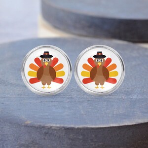 May include: A pair of round stud earrings featuring a graphic illustration of a brown turkey wearing a black pilgrim hat. The turkey has a fanned tail with alternating red, orange, and yellow feathers, a yellow beak, and small feet set in a silver frame.