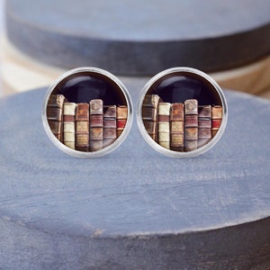 Peut inclure: Une paire de boutons de manchette ronds en argent. Chaque bouton de manchette présente une image miniature d'une rangée de livres anciens aux couvertures usagées. Les livres sont de couleurs variées, beige, marron et rouge, sur fond sombre.
