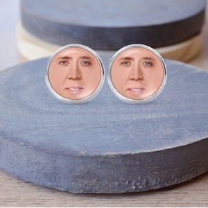 Op de afbeelding: Een paar zilverkleurige oorbellen met ronde, gefacetteerde glazen cabochons met een close-up afbeelding van het gezicht van Nicolas Cage.