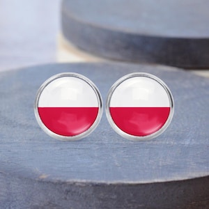 Może przedstawiać: Para okrągłych kolczyków sztyftów z motywem flagi Polski. Każdy kolczyk ma białą górną połowę i czerwoną dolną połowę, osadzone w srebrnej oprawie. Kolczyki są prezentowane na niebieskiej powierzchni.