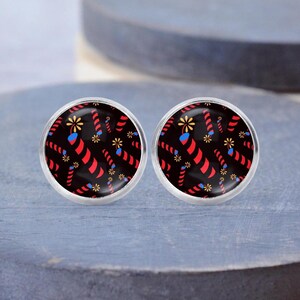 Puede incluir: Un par de pendientes redondos plateados. Cada pendiente presenta un fondo negro con un patrón repetido de fuegos artificiales rojos y amarillos y detalles azules. Los pendientes se muestran sobre una superficie azul.