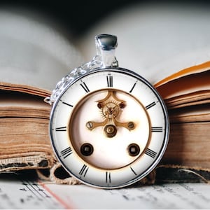 Könnte beinhalten: Eine silberne Halskette mit einem weißen Zifferblatt und sichtbaren Zahnrädern. Das Zifferblatt hat römische Ziffern und einen goldenen Rand. Die Halskette liegt auf einem geöffneten Buch mit alten Seiten.