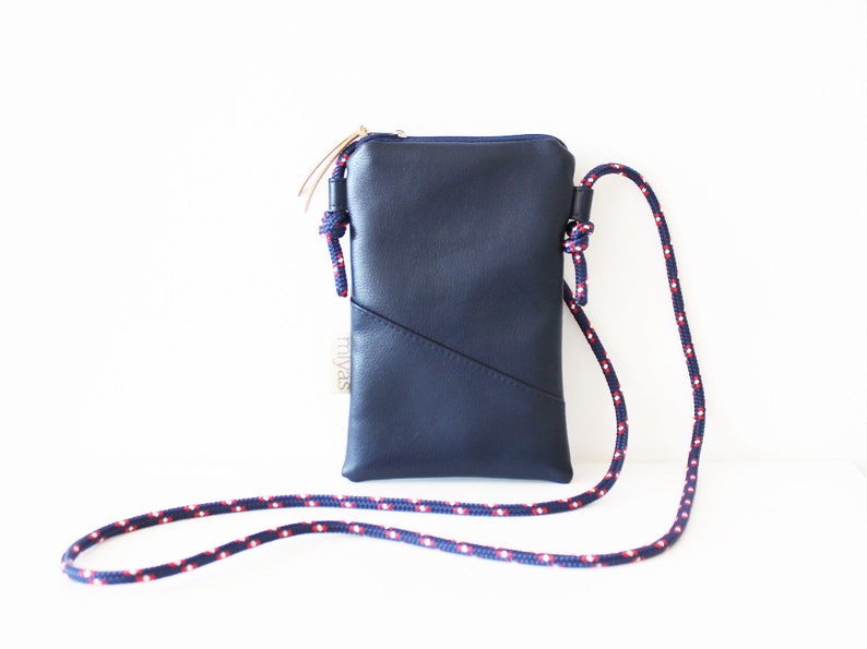 May include: A small navy blue leather crossbody bag with a zipper closure and a red and white rope strap. The bag has a small label that says "Truvas".