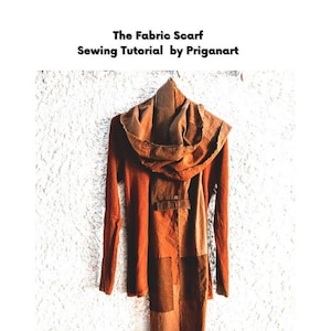 Peut inclure: Une écharpe en tissu marron avec un motif patchwork, suspendue à un cintre contre un mur texturé blanc. L'écharpe est faite de différents tissus marrons cousus ensemble. Le texte "The Fabric Scarf Sewing Tutorial by Priganart" est en haut de l'image.