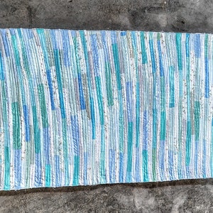 Puede incluir: Una alfombra hecha a mano con un patr&oacute;n de rayas en tonos azules, verdes y blancos. La alfombra est&aacute; hecha de recortes de tela reciclados y tiene un aspecto r&uacute;stico y hecho a mano.