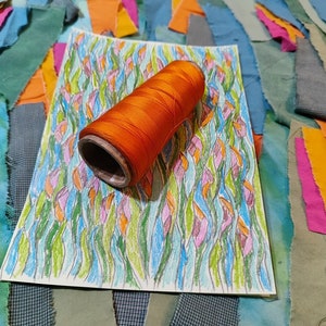May include: A spool of orange thread on a white sheet of paper with a colourful drawing of stylised flowers. The paper is surrounded by scraps of fabric in various shades of blue, green, orange, and pink.