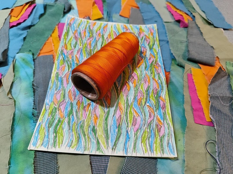 May include: A spool of orange thread on a white sheet of paper with a colourful drawing of stylised flowers. The paper is surrounded by scraps of fabric in various shades of blue, green, orange, and pink.