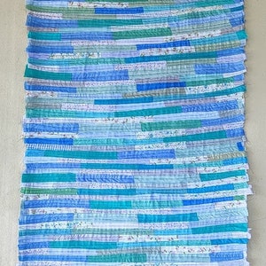 Puede incluir: Una colcha hecha a mano con tiras de tela en tonos azules, verdes y blancos. La colcha est&aacute; colgada de una percha y tiene un aspecto r&uacute;stico y vintage.