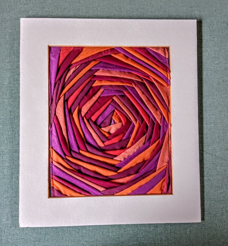 Pu&ograve; includere: Un'opera d'arte incorniciata che presenta un motivo a spirale di tessuto in tonalit&agrave; di arancione, rosa e viola.